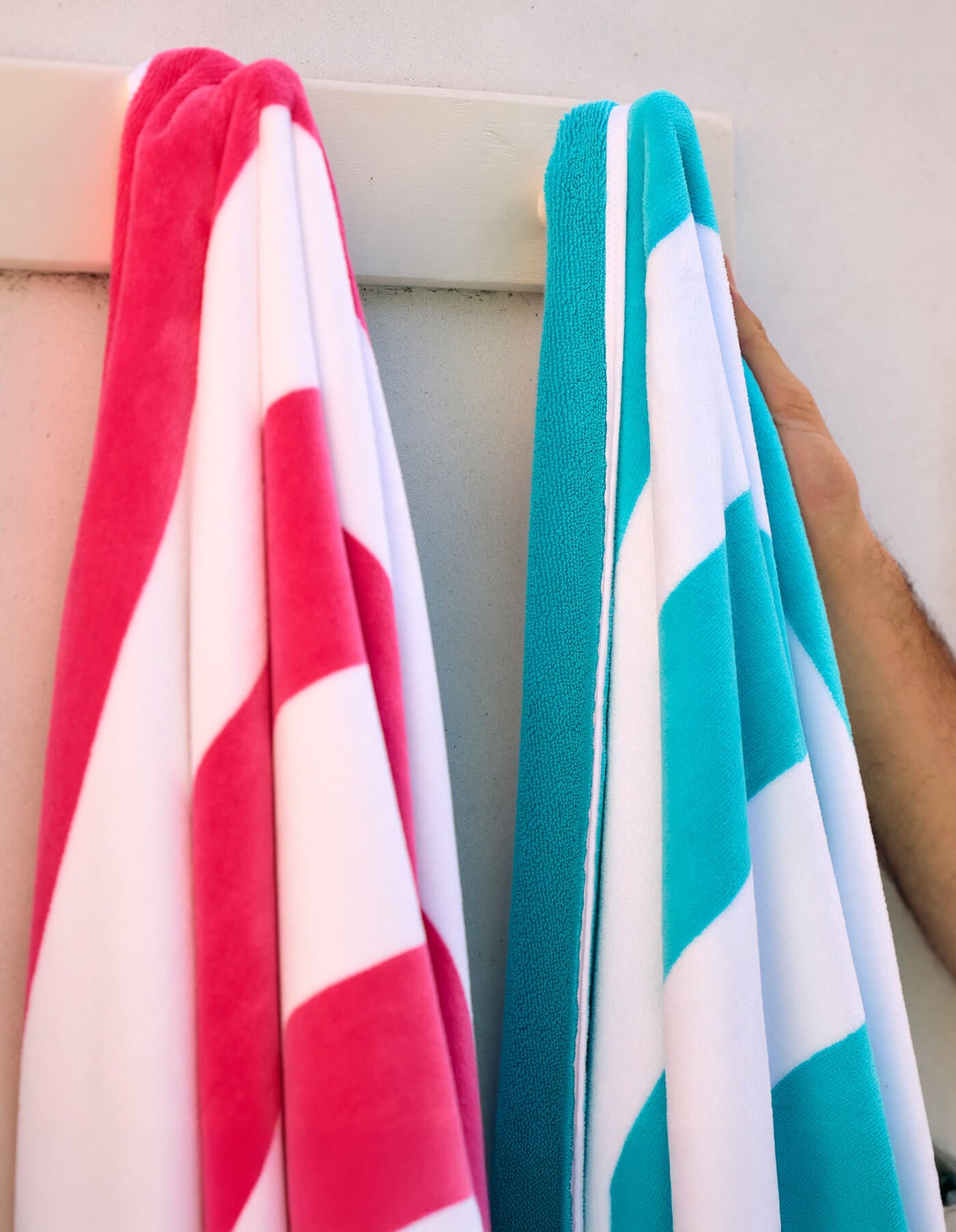 Two Cozy Earth Seaside Resort Towels, one pink and white and the other blue and white, hang on a white rack against a light wall. A person’s arm is holding the blue towel.