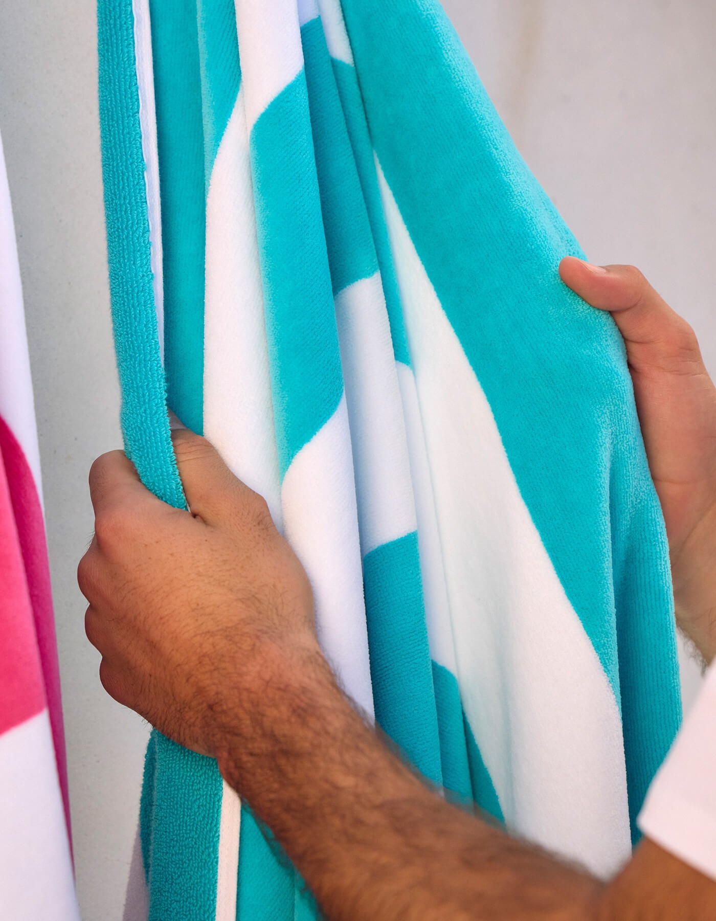A close-up of two hands holding the Cozy Earth Seaside Resort Towel in blue and white stripes, with part of a pink and white version beside it. The background is plain and blurred.