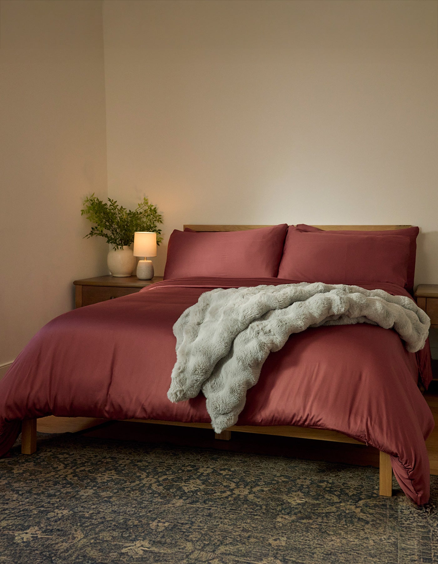 A neatly made bed features Cozy Earth's Bamboo Duvet Cover in dark red and a soft gray throw, set against a plain wall. A wooden nightstand with a plant, lamp, and candle sits beside the bed, with a patterned rug on the floor.