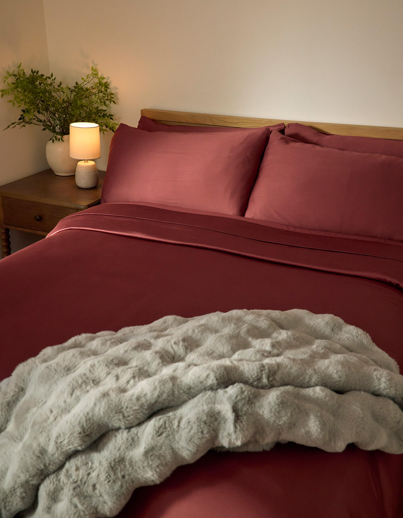 A neatly made bed featuring Cozy Earth’s Bamboo Sheet Set in deep red, paired with a light gray textured blanket, a wooden nightstand with a lamp and green plant, all set against a plain cream wall.