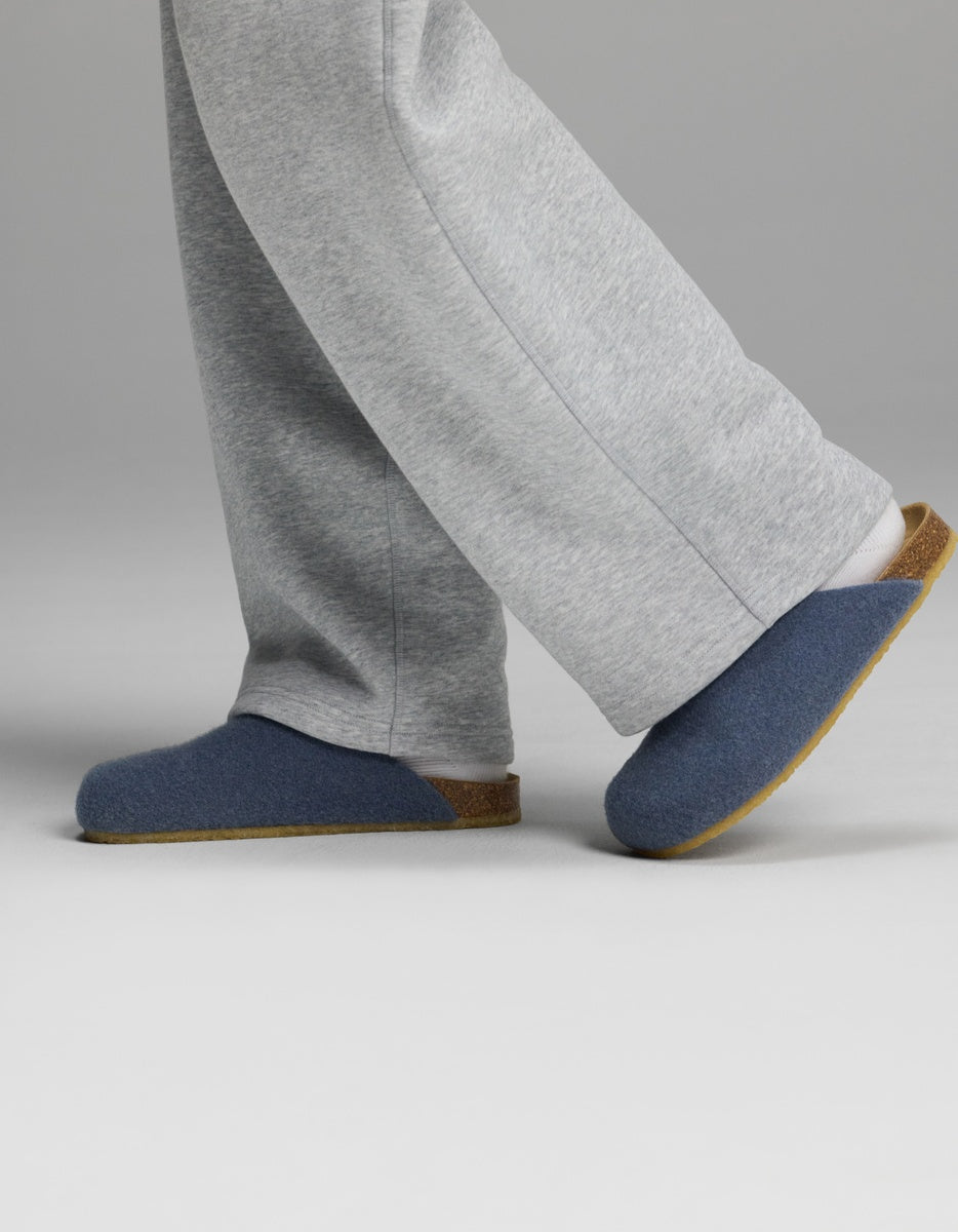 A person wearing grey sweatpants and Cozy Earth Lakehouse Clog slippers, featuring blue uppers and brown soles, walking on a plain light background.