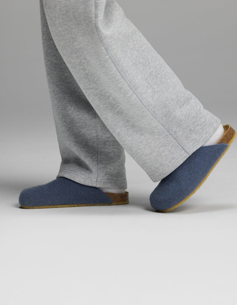 A person wearing grey sweatpants and Cozy Earth Lakehouse Clog slippers, featuring blue uppers and brown soles, walking on a plain light background.