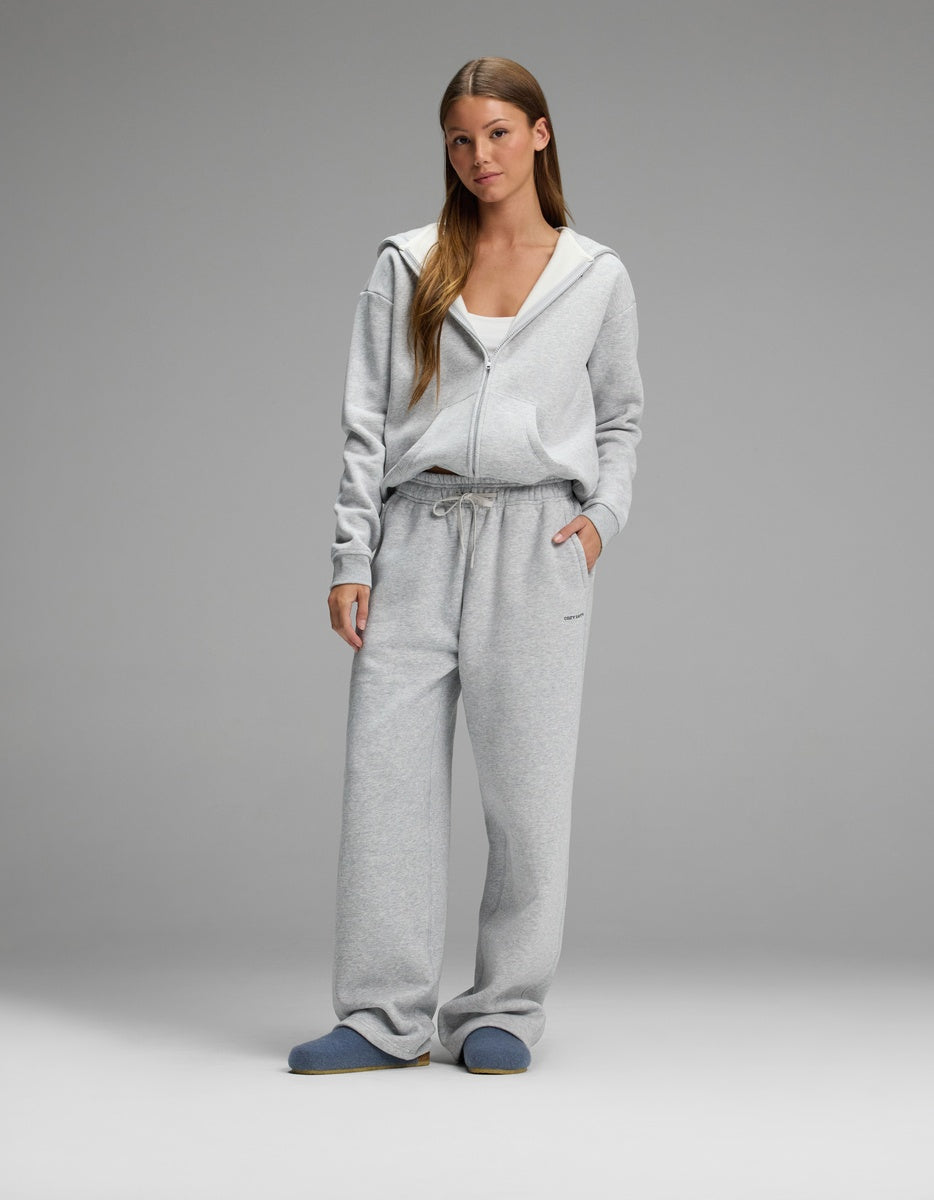 A woman stands against a plain background wearing a light gray zip-up hoodie, matching gray sweatpants, a white top underneath, and Cozy Earth Lakehouse Clog slippers in blue. She has long hair and her hands are in her pockets.