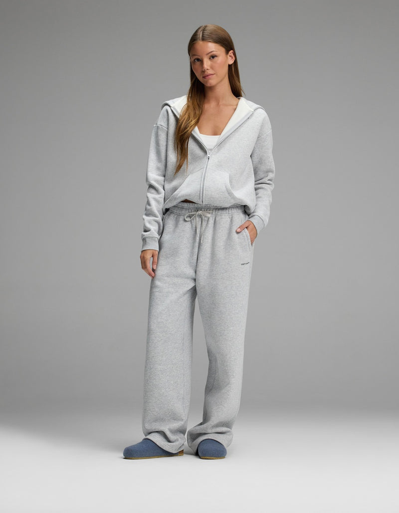 A woman stands against a plain background wearing a light gray zip-up hoodie, matching gray sweatpants, a white top underneath, and Cozy Earth Lakehouse Clog slippers in blue. She has long hair and her hands are in her pockets.