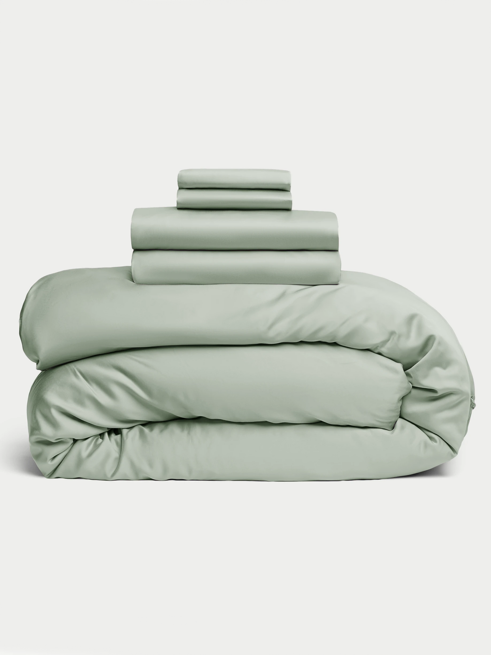 The Bamboo Bedding Deluxe Bundle from Cozy Earth is neatly stacked in pastel green, with folded sheets and pillowcases atop a fluffy duvet. A plain white background highlights the bedding's soft texture and gentle color. 