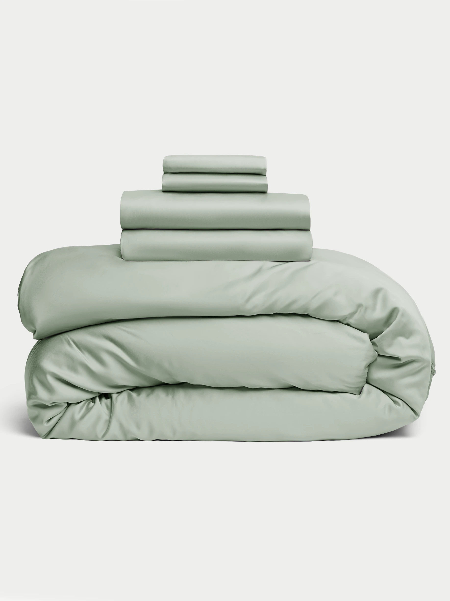 The Bamboo Bedding Deluxe Bundle from Cozy Earth is neatly stacked in pastel green, with folded sheets and pillowcases atop a fluffy duvet. A plain white background highlights the bedding's soft texture and gentle color. |Color:Laurel