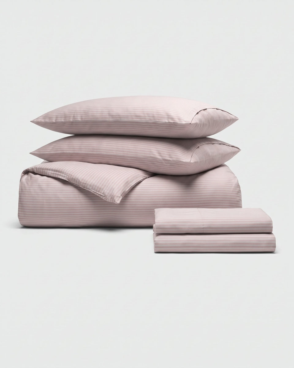 A Cozy Earth Bamboo Bedding Deluxe Bundle in cream, featuring a lavender striped pattern, with two pillows stacked atop a folded duvet and neatly folded sheets, displayed on a plain white background.