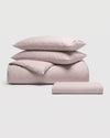 The Cozy Earth Bamboo Bedding Essential Bundle is displayed on a plain white background, featuring a neatly stacked set of light mauve bedding that includes two pillows, a folded sheet, and a rolled sheet.|Color:Classic Stripe in Lavender