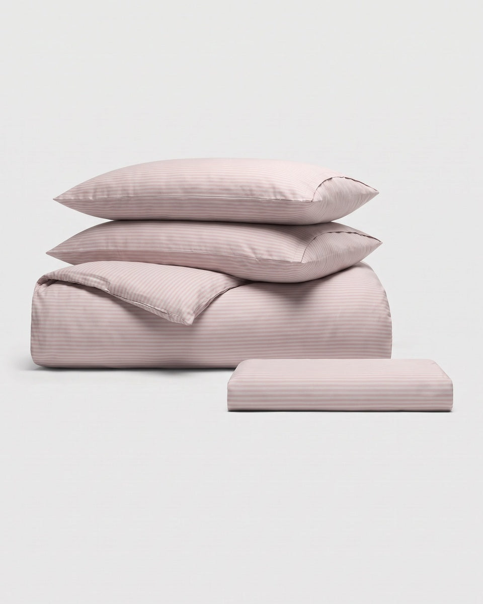 The Cozy Earth Bamboo Bedding Essential Bundle is displayed on a plain white background, featuring a neatly stacked set of light mauve bedding that includes two pillows, a folded sheet, and a rolled sheet.