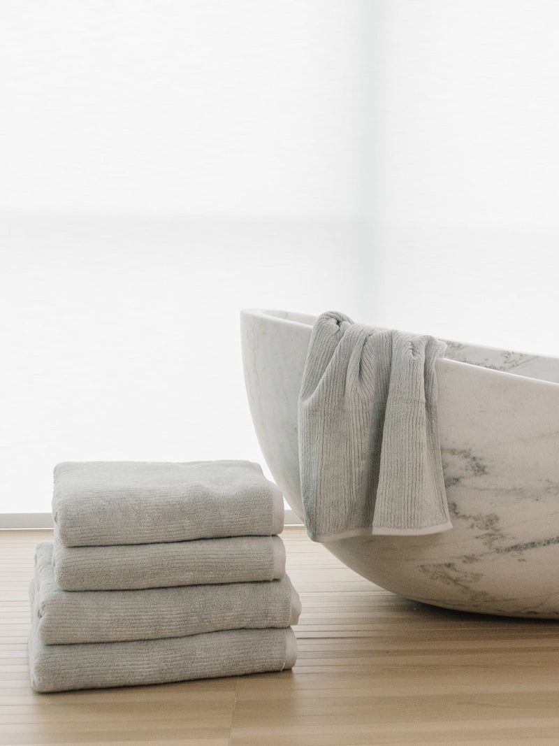 The Complete Ribbed Terry Bath Bundle by Cozy Earth features four neatly folded gray towels stacked next to a white marble bathtub, with another towel draped over the edge, all on a light wooden floor in a bright, minimalist setting.
