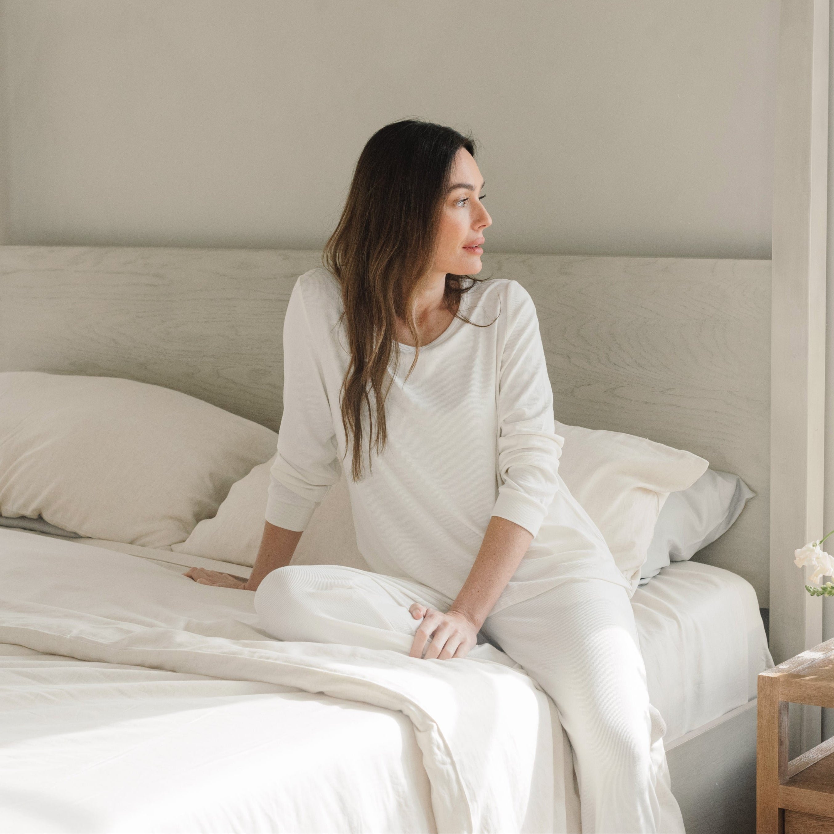 Woman in white pajamas sits on a bed with white sheets, looking to the side. The room has neutral tones and a flower in a small vase on the bedside table.