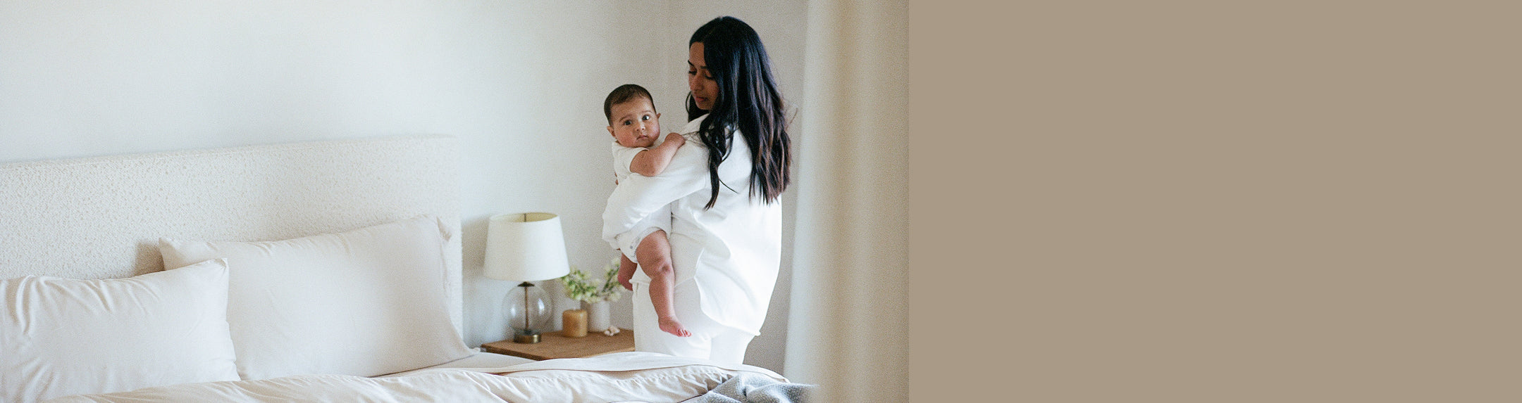 Woman holding a baby in a bedroom with a white couch and lamp.