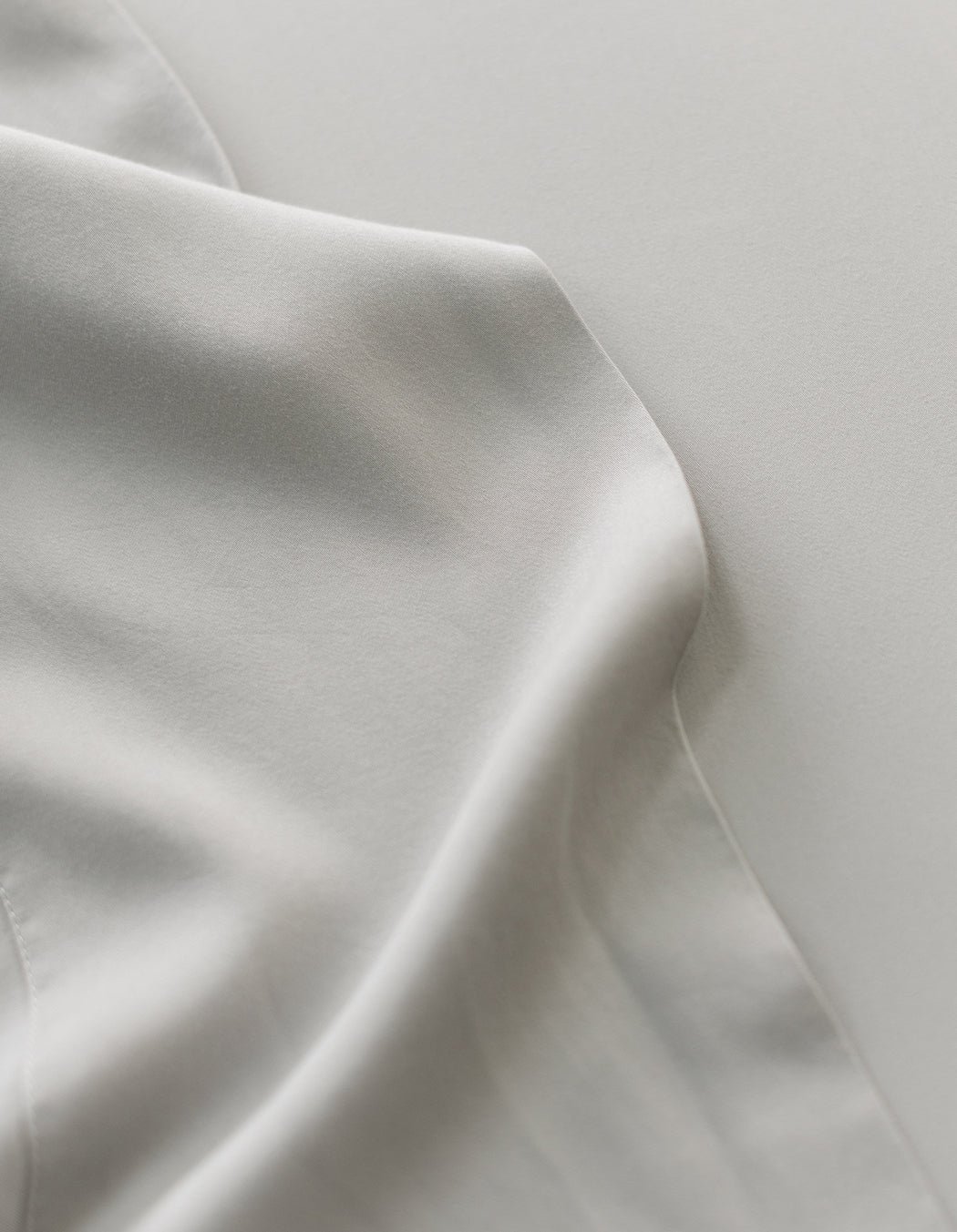 A close-up of Cozy Earth's Bamboo Sheet Set showcases smooth, light grey fabric with gentle folds creating soft shadows. The silky, slightly shiny texture adds elegance to the image. 