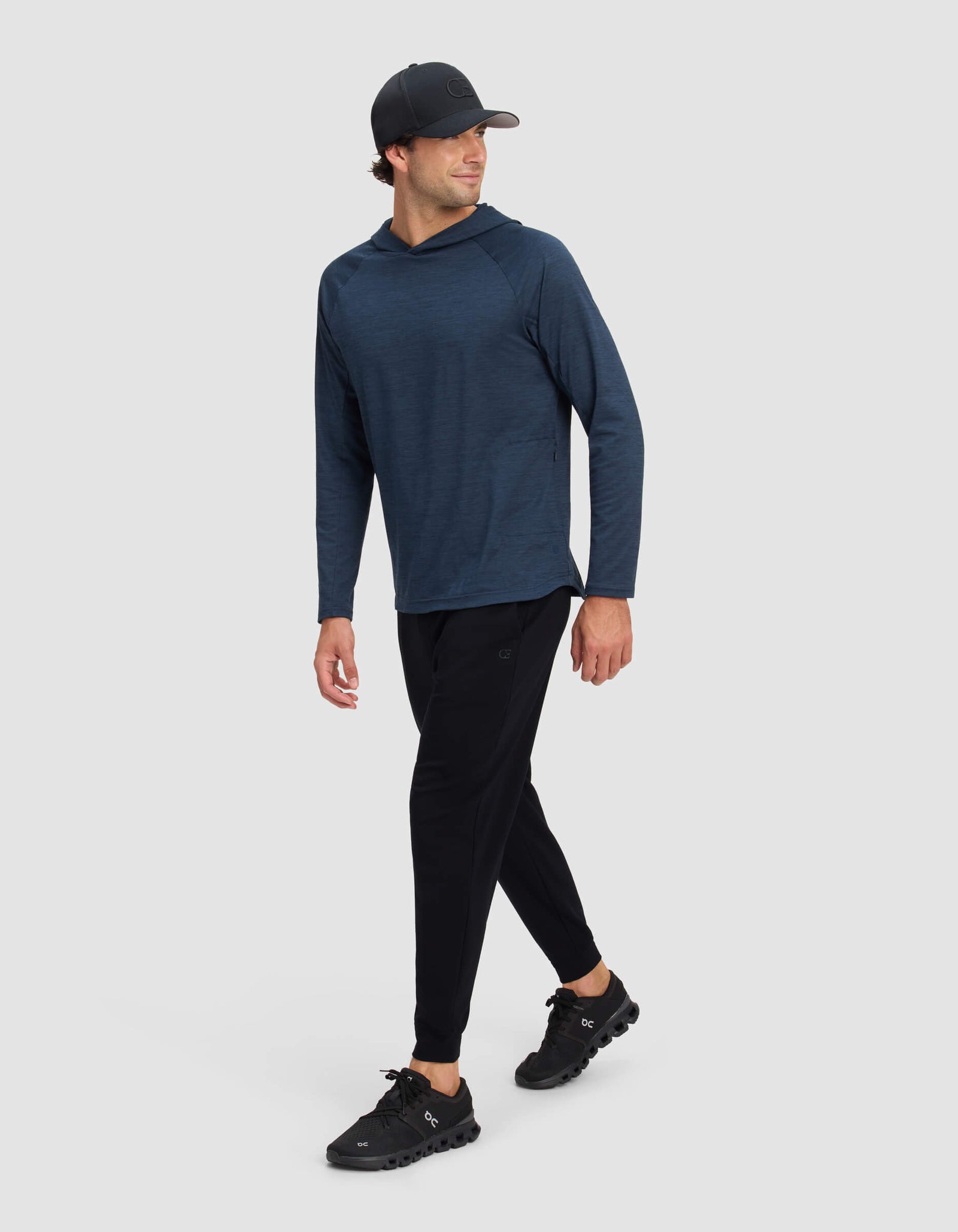 A man in a Cozy Earth Men's Lightweight Performance Hoodie, dark baseball cap, black jogger pants, and black athletic shoes walks confidently against a plain light gray background.