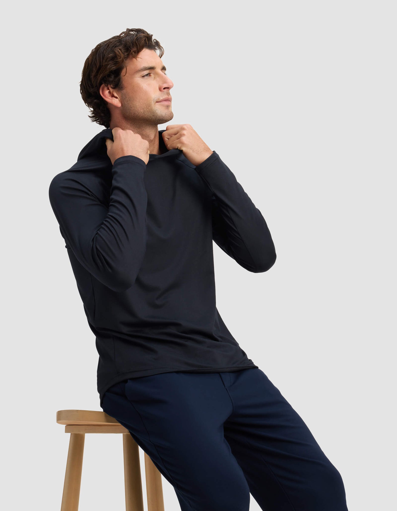 A man wears the Cozy Earth Men's Lightweight Performance Hoodie in black with navy pants, sitting on a wooden stool and adjusting his hoodie while looking slightly upward against a plain light background.