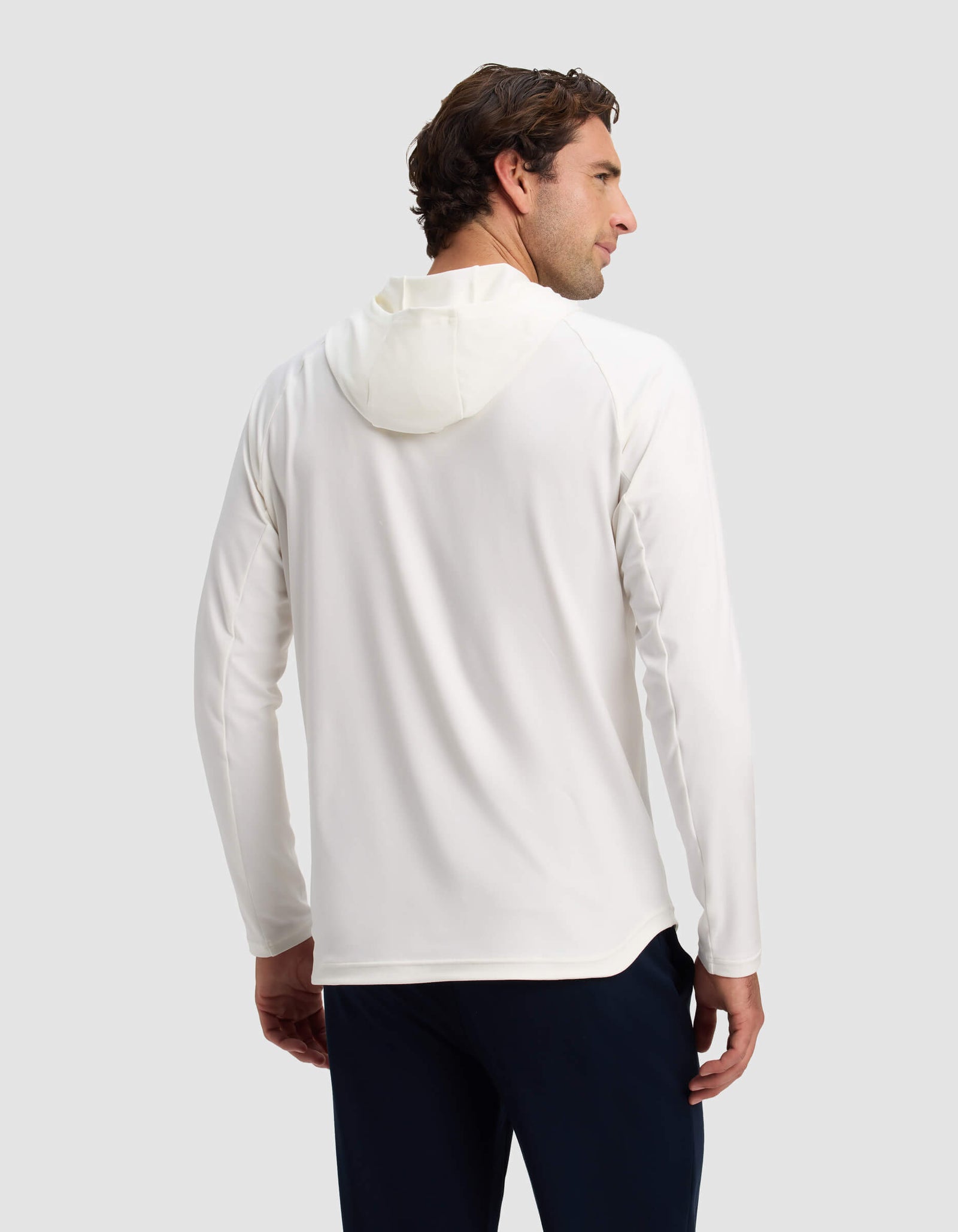 A man in the Cozy Earth Men's Lightweight Performance Hoodie, a white long-sleeve top, and dark pants stands facing away and looks over his left shoulder against a plain light gray background.