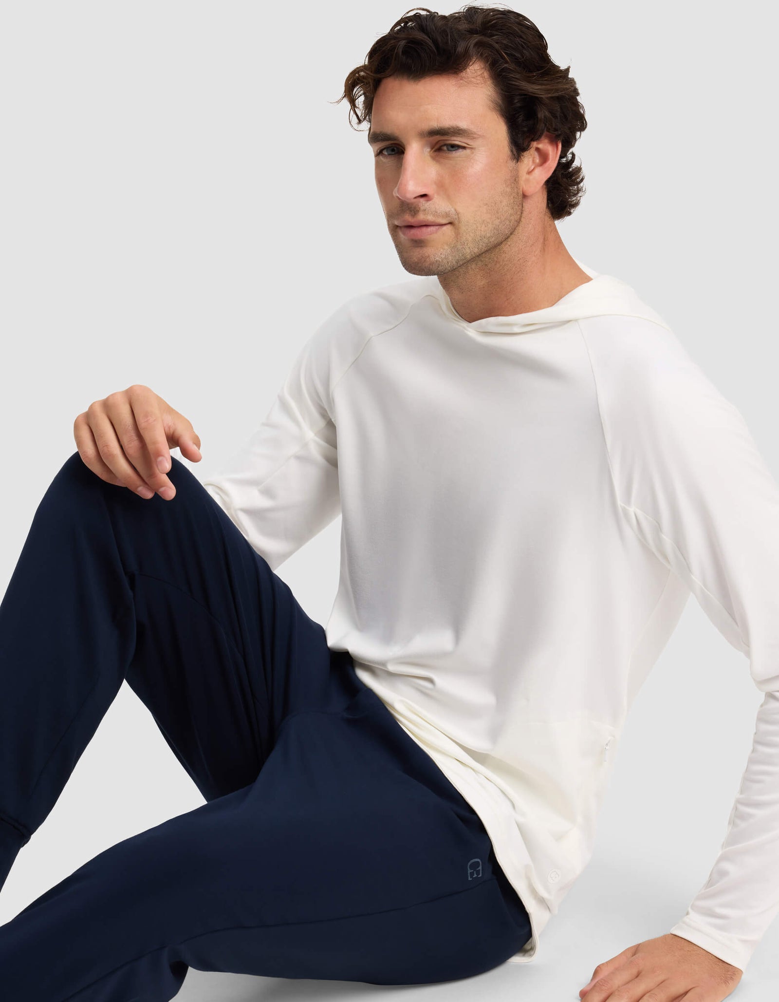 A man with wavy brown hair sits on the floor in a long-sleeve white Men's Lightweight Performance Hoodie by Cozy Earth and dark blue pants. He looks at the camera with a neutral expression, against a plain light gray background.
