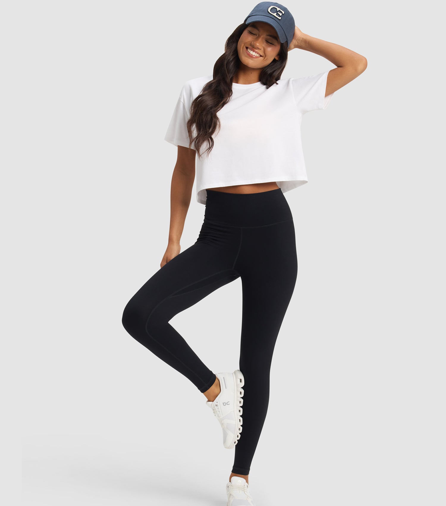 Woman wearing a white crop top, black leggings, and a blue cap on a light gray background
