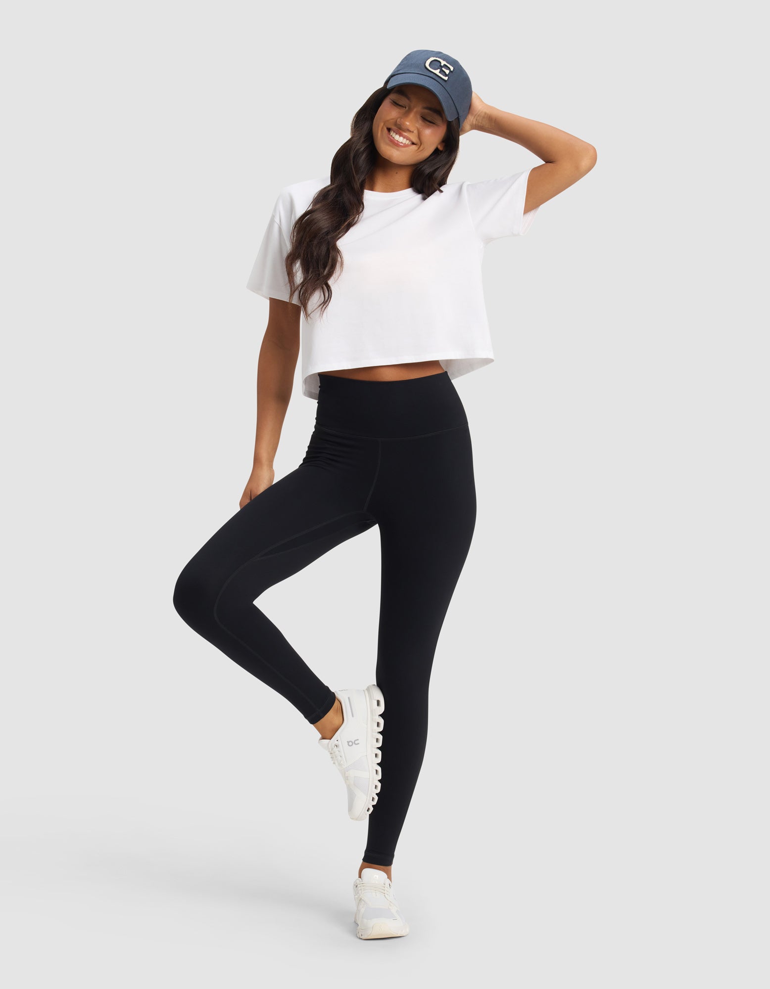 A woman smiles against a light gray background, wearing Cozy Earth's Women's Liv Legging, a white cropped t-shirt, white sneakers, and a blue baseball cap, with one hand on her head and one leg slightly raised.