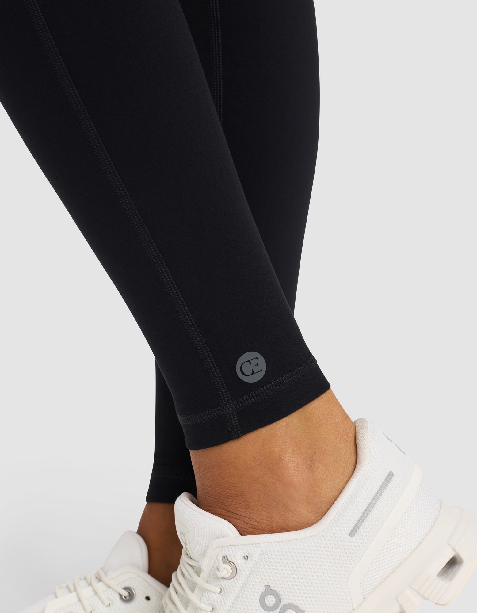 Close-up of a person wearing Cozy Earth Women's Liv Legging in black with a small circular logo near the ankle, paired with white athletic shoes. The background is plain and light-colored.