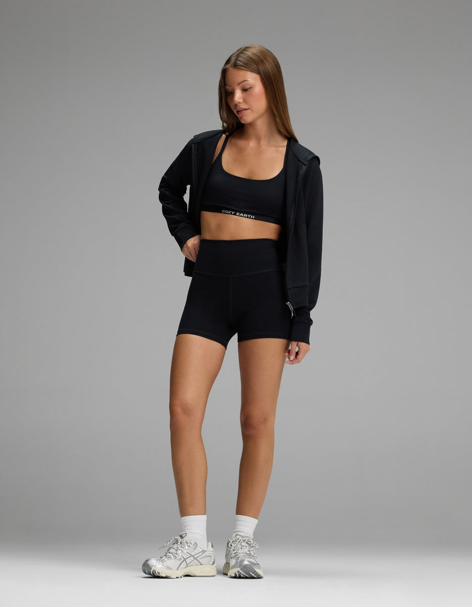 A woman wears a black sports bra, black Women’s Liv Biker Short 4″ by Cozy Earth, and a black zip-up hoodie with white sneakers.