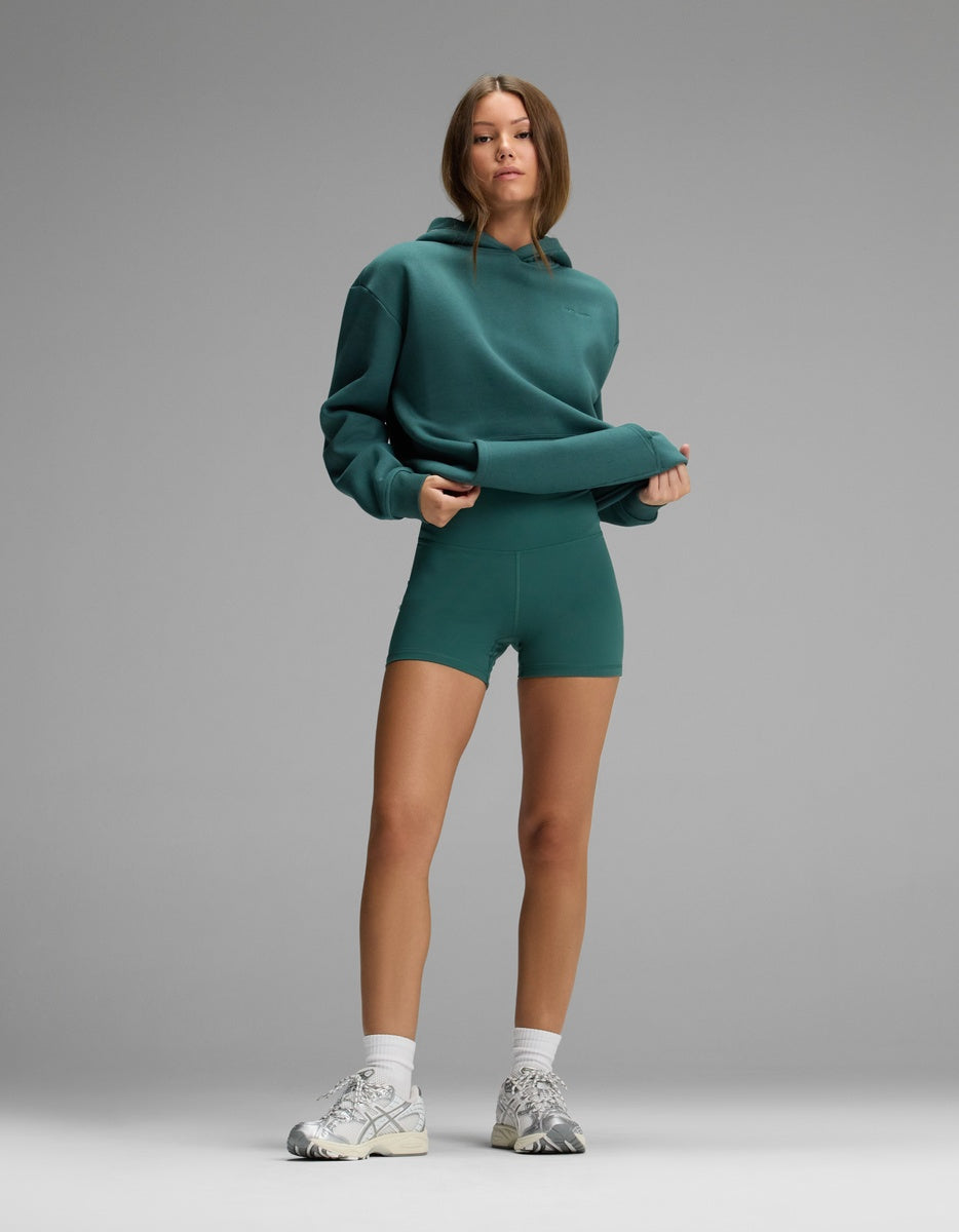 A person wears a teal hooded sweatshirt and matching Women’s Liv Biker Short 4″ with white socks and athletic sneakers.
