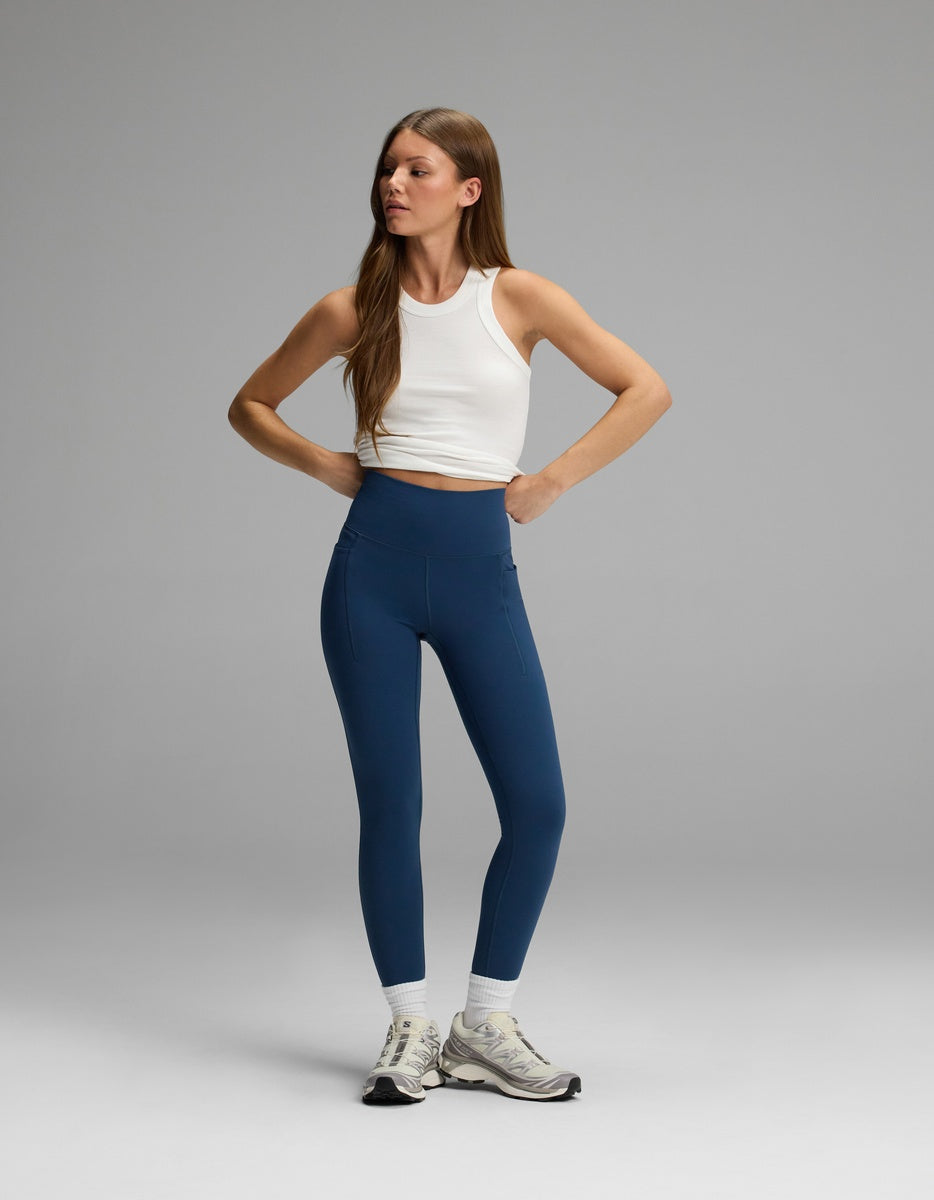 A woman wearing a white tank top and blue Women’s Liv Leggings with Pockets that feature a high-waisted fit.
