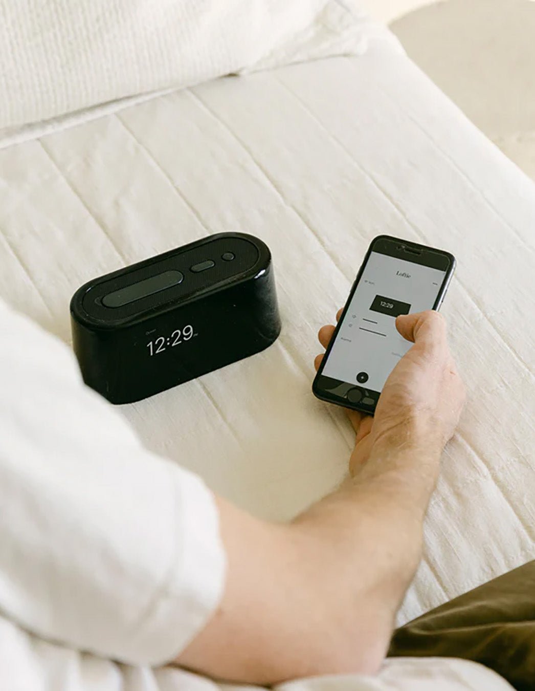 A person on a Bamboo Sheet Set controls the Cozy Earth Loftie Clock (showing 12:29) via smartphone, with both devices on a white bedsheet.