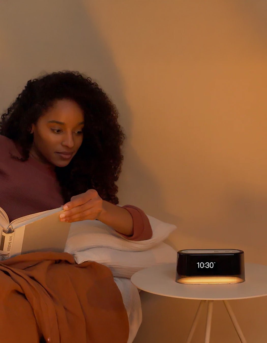 A woman relaxes in bed with a book, accompanied by the Cozy Earth Bamboo Sheet Set and Loftie Clock on a round white bedside table under warm lighting.