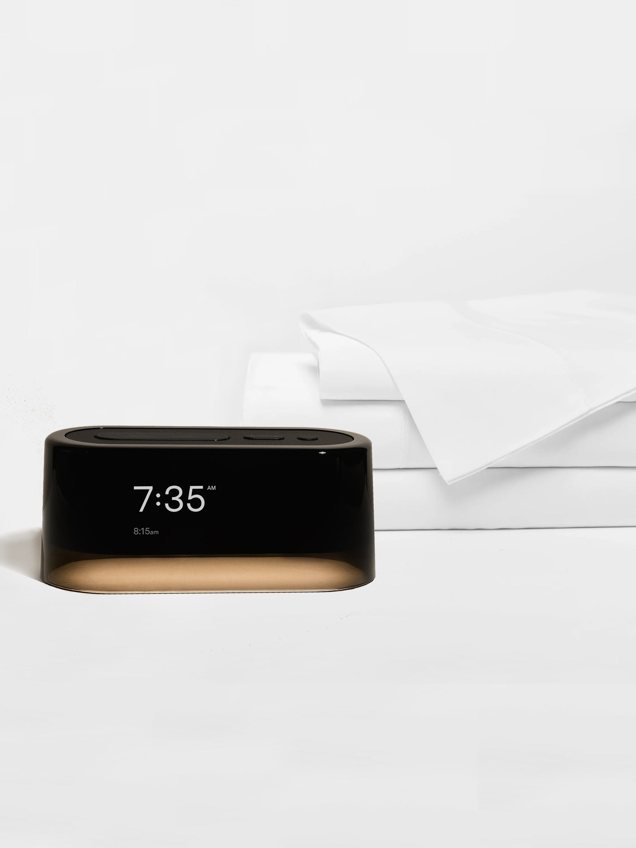 The Cozy Earth Bamboo Sheet Set x Loftie Clock features a sleek black digital alarm clock showing 7:35 AM on a white surface, next to neatly folded white bamboo sheets against a minimal white background.