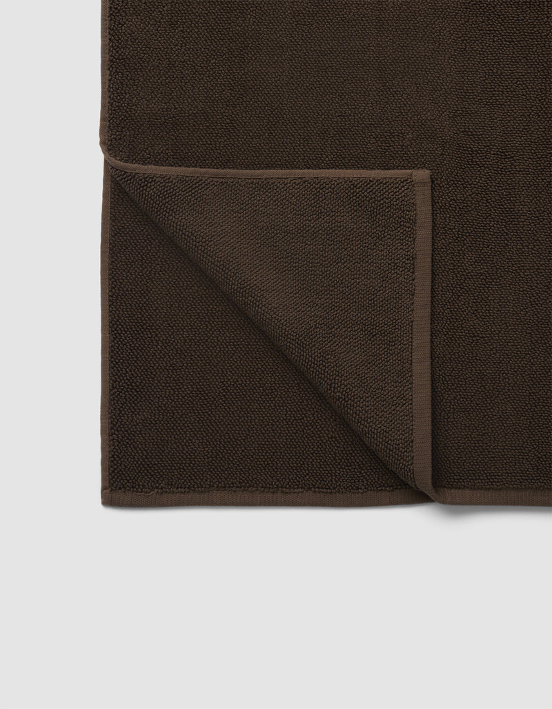 The Cozy Earth Loop Terry Bath Mat, shown in dark brown with a folded corner revealing the reverse side, is displayed against a plain white background.