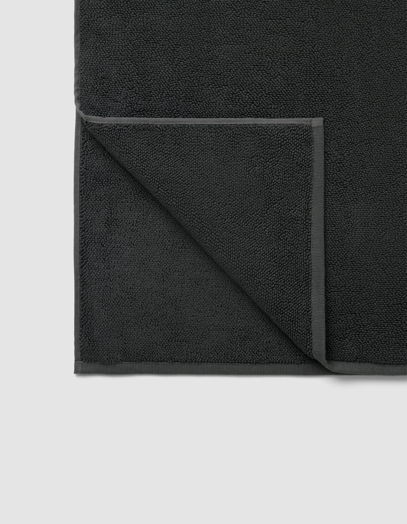 The Cozy Earth Loop Terry Bath Mat in dark gray is pictured neatly folded at one corner, displaying its textured surface and smooth edge border on both front and back sides.