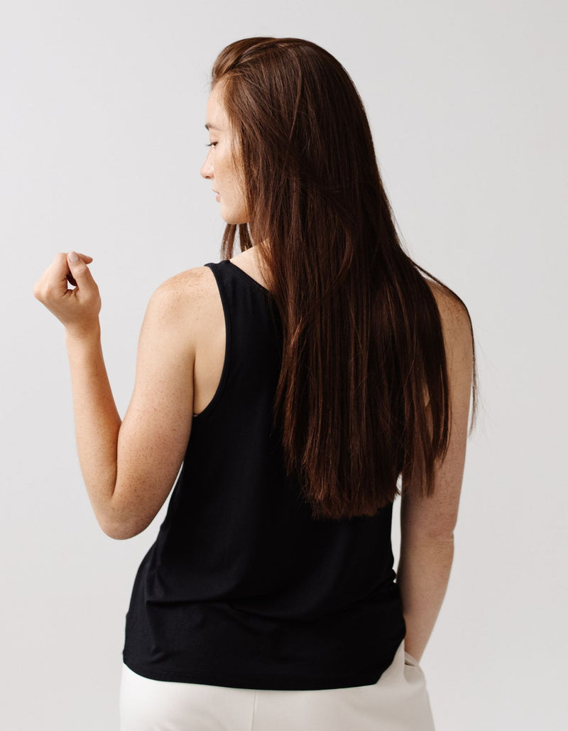A woman with long brown hair models the Cozy Earth Women's Bamboo Stretch-Knit Lounge Tank, a sleeveless black top, paired with white pants. She stands against a plain light background, facing away with her head turned slightly to the side.