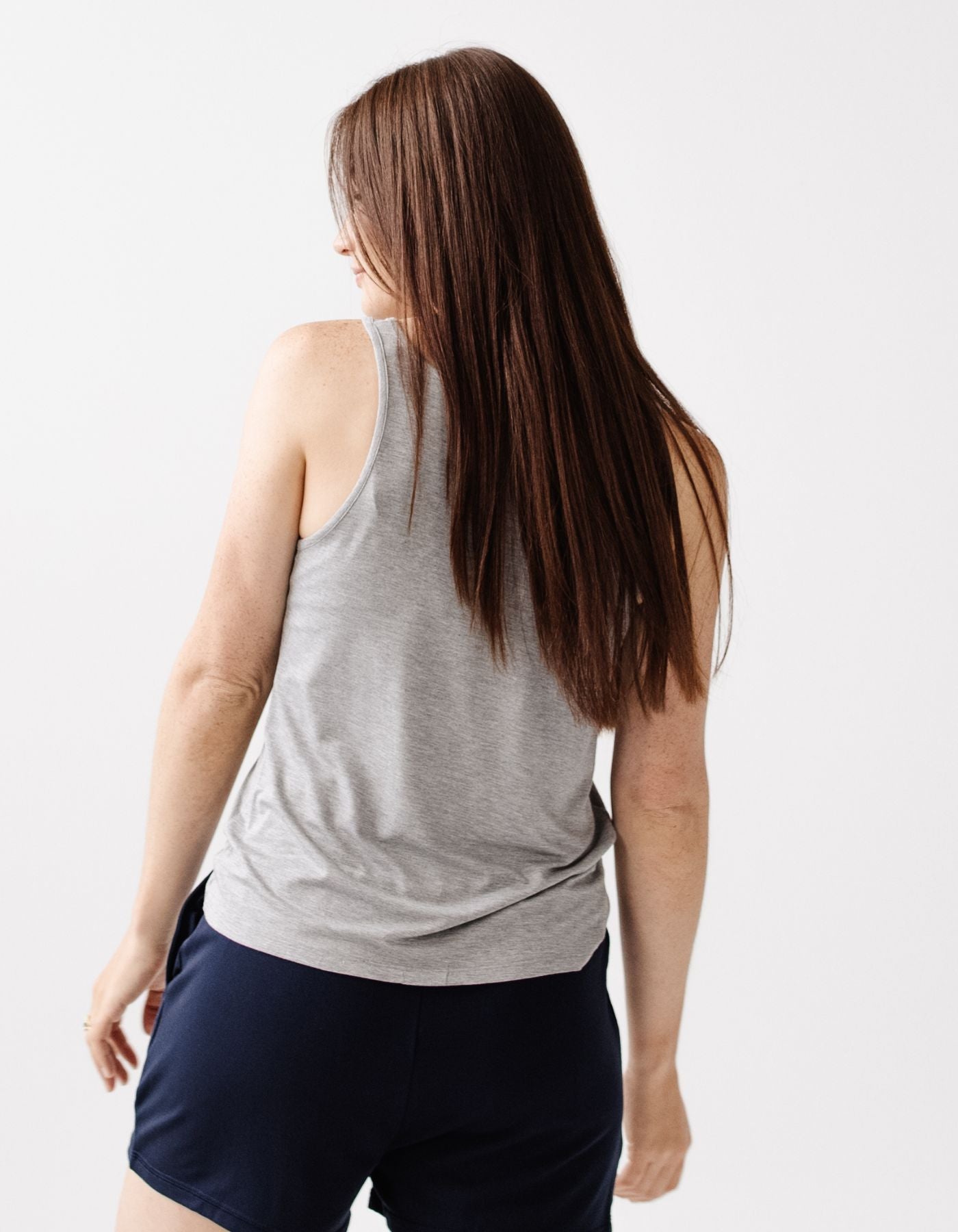 A woman with long brown hair wears the Cozy Earth Women's Bamboo Stretch-Knit Lounge Tank in gray, paired with navy shorts, standing against a plain white background and looking slightly over her shoulder.