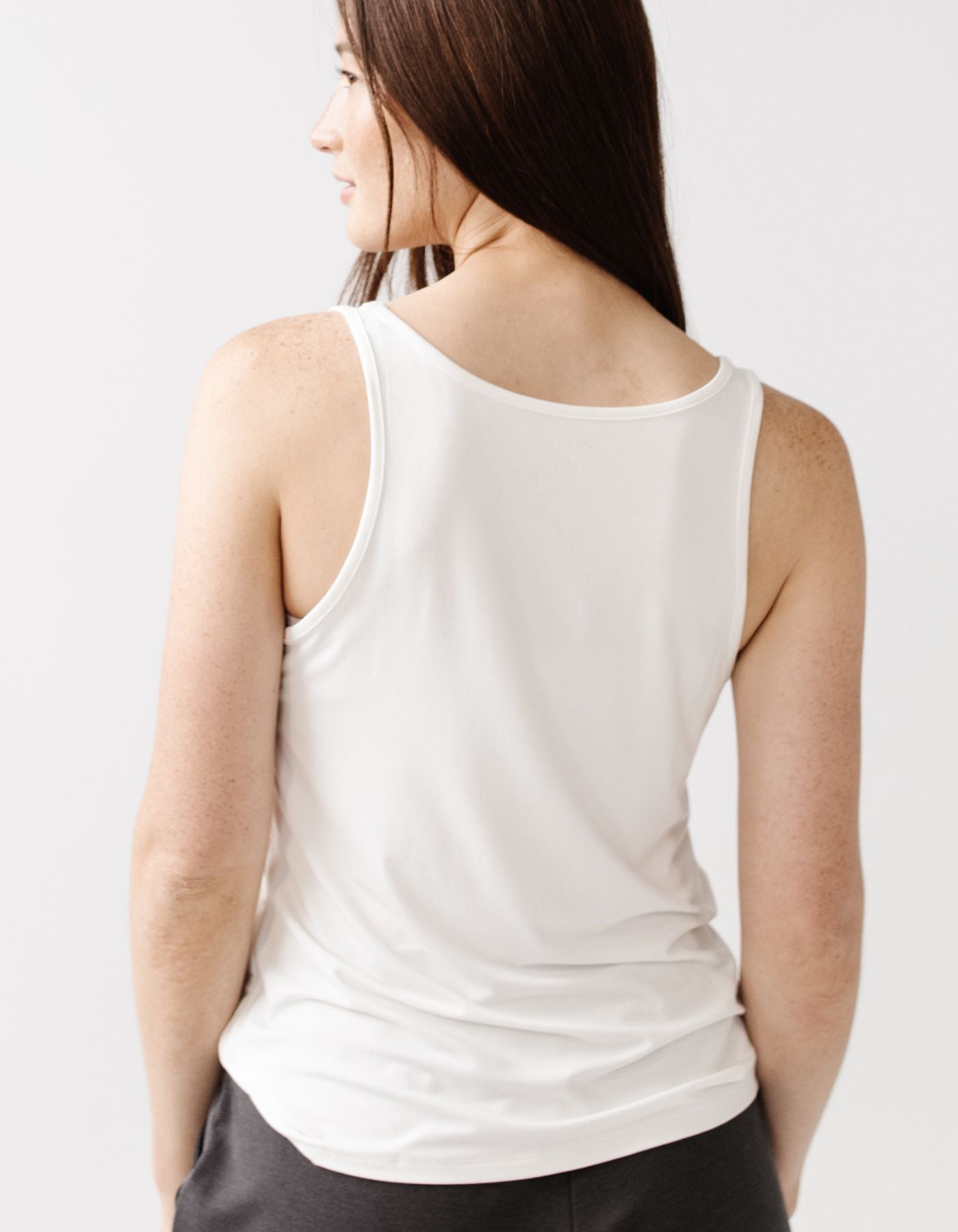 A woman with long brown hair is seen from behind wearing the Cozy Earth Women's Bamboo Stretch-Knit Lounge Tank in white, paired with dark pants, standing before a plain light background.