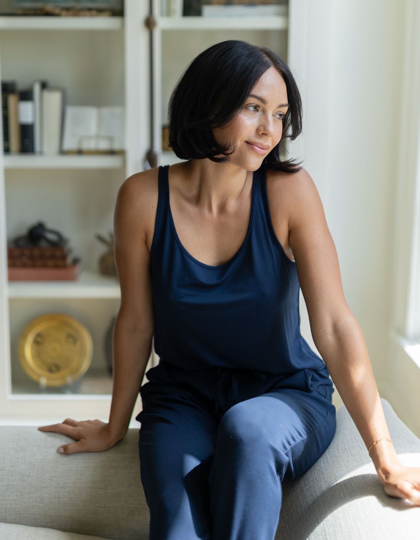 A woman with short dark hair, wearing a navy Women's Bamboo Stretch-Knit Lounge Tank by Cozy Earth, sits on a light couch near a window, smiling and looking to the side. Bookshelves appear in the background.