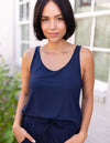 A woman with short dark hair stands outside, smiling softly with her hands in her pockets, wearing Cozy Earth's Women's Bamboo Stretch-Knit Lounge Tank in navy. Greenery and windows are visible in the background.|Color:Navy