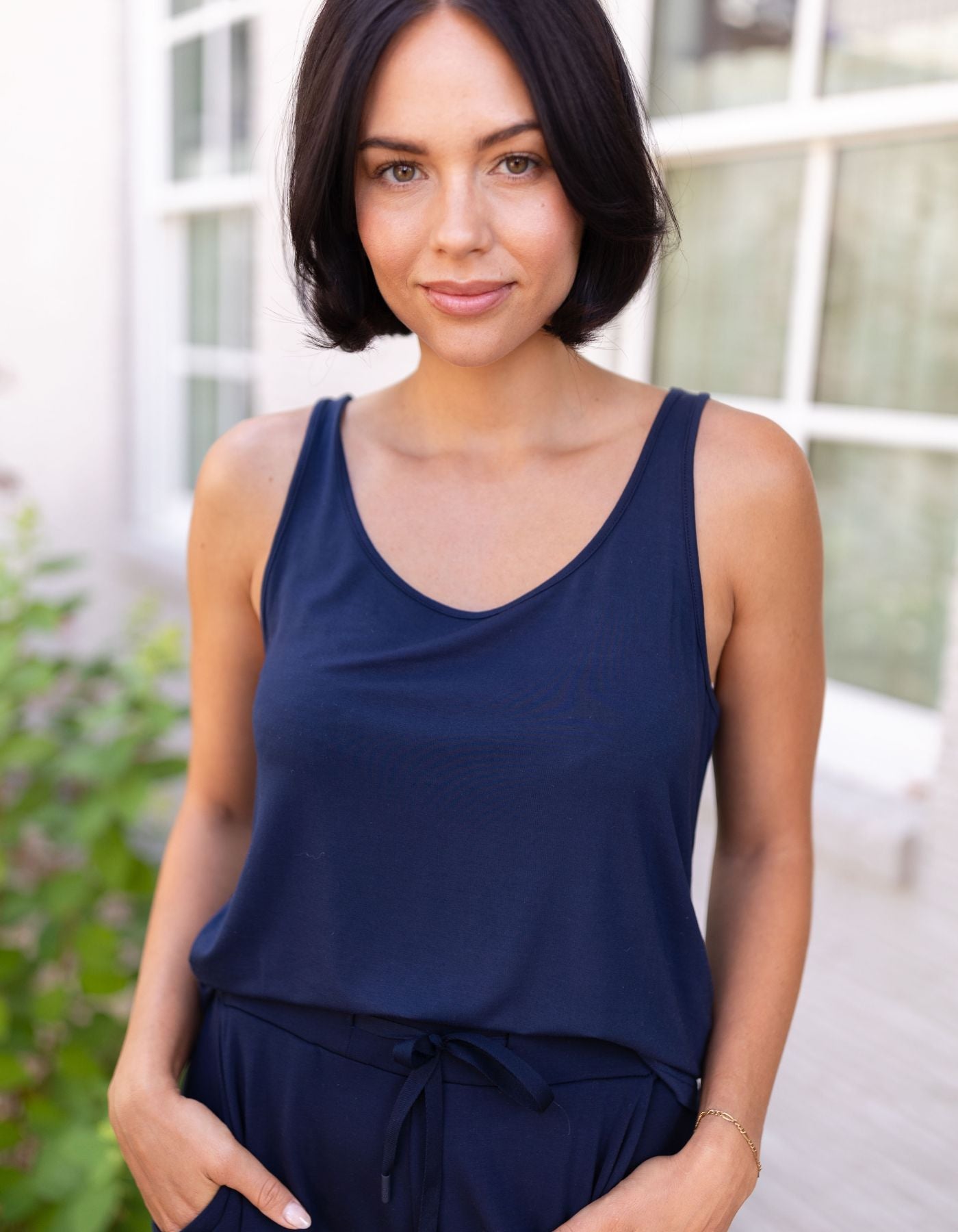 A woman with short dark hair stands outside, smiling softly with her hands in her pockets, wearing Cozy Earth's Women's Bamboo Stretch-Knit Lounge Tank in navy. Greenery and windows are visible in the background.|Color:Navy