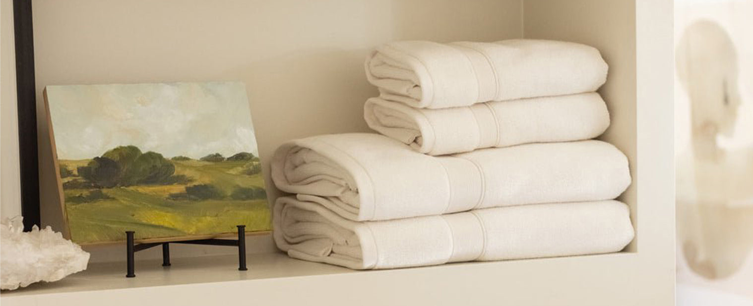 Stack of white Cozy Earth Luxe Bathtowels on a shelf next to a small painting of a landscape
