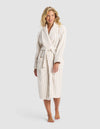 A smiling woman with wavy blonde hair models the Cozy Earth Striped Luxe Bath Robe, featuring beige vertical stripes, a tie belt, and large front pockets. She stands barefoot against a light gray background. |Color:Sand Stripe