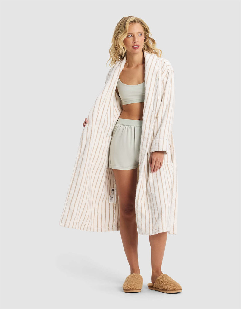 A woman stands against a plain light gray background, wearing the Cozy Earth Striped Luxe Bath Robe over a light green bralette and shorts set, paired with tan slippers.