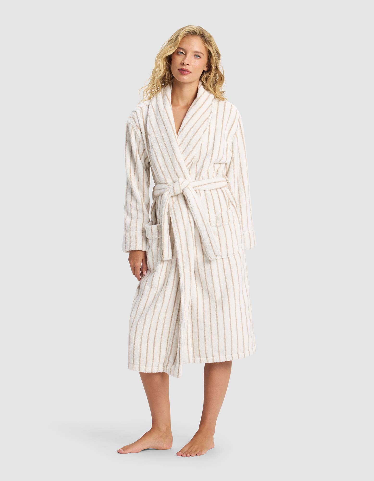 A woman stands barefoot against a light gray background, wearing the Cozy Earth Striped Luxe Bath Robe—white with beige vertical stripes and a belted waist.|Color:Sand Stripe