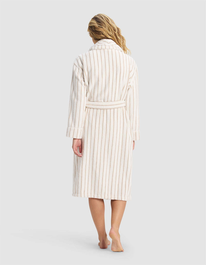 A woman with wavy blonde hair stands barefoot on a plain light background, shown from the back in Cozy Earth's Striped Luxe Bath Robe—a long, cream robe with beige vertical stripes and a belt.
