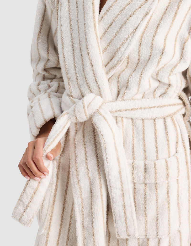 A person wears the Cozy Earth Striped Luxe Bath Robe, a soft cream-colored robe with beige vertical stripes, tied at the waist; only their upper body and one hand adjusting the belt are visible.