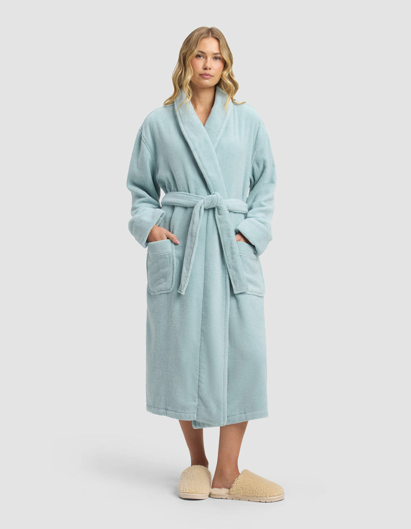 A woman stands facing forward wearing the Cozy Earth Luxe Bath Robe—light blue with a belt and large pockets—and beige slippers, set against a plain light background.