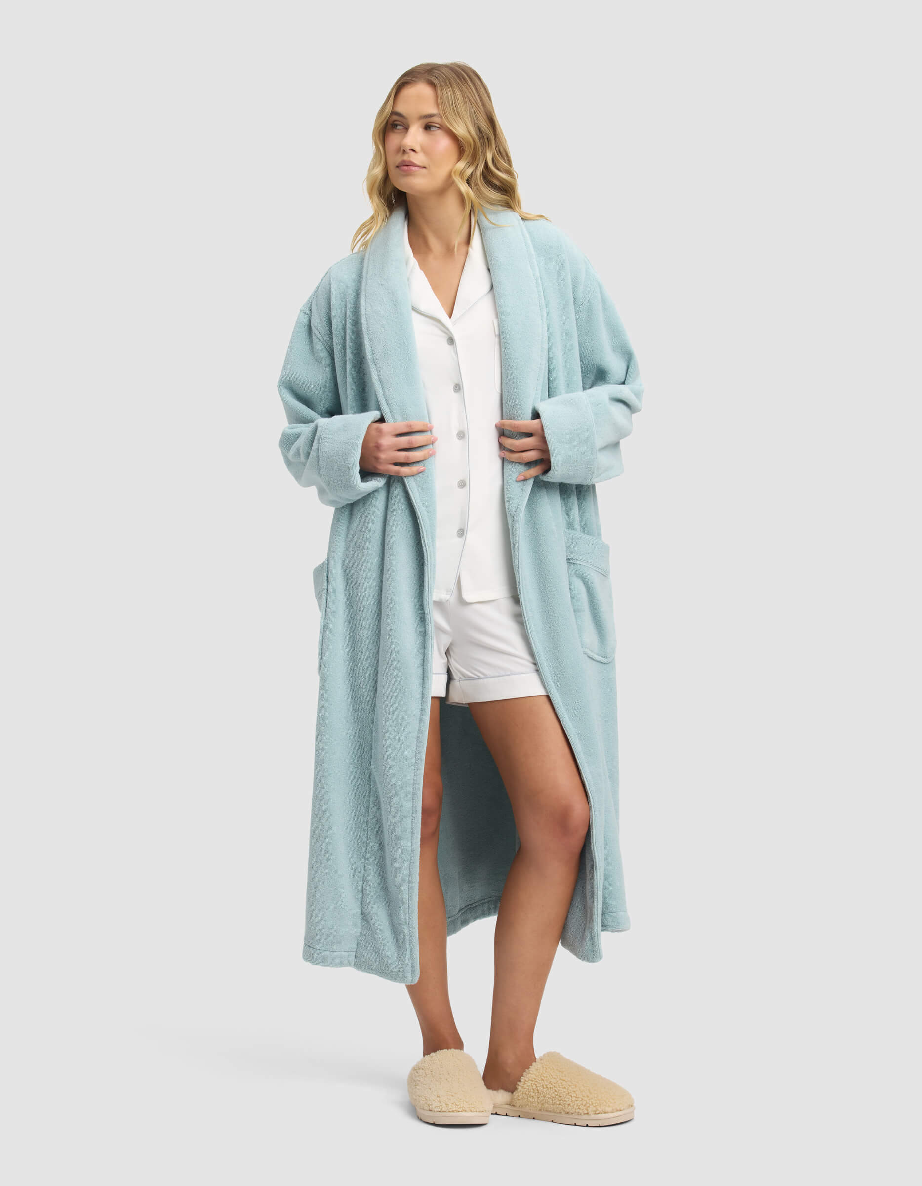 A woman with wavy blonde hair wears the Cozy Earth Luxe Bath Robe over white pajamas and beige slippers, looking relaxed against a plain light gray background.