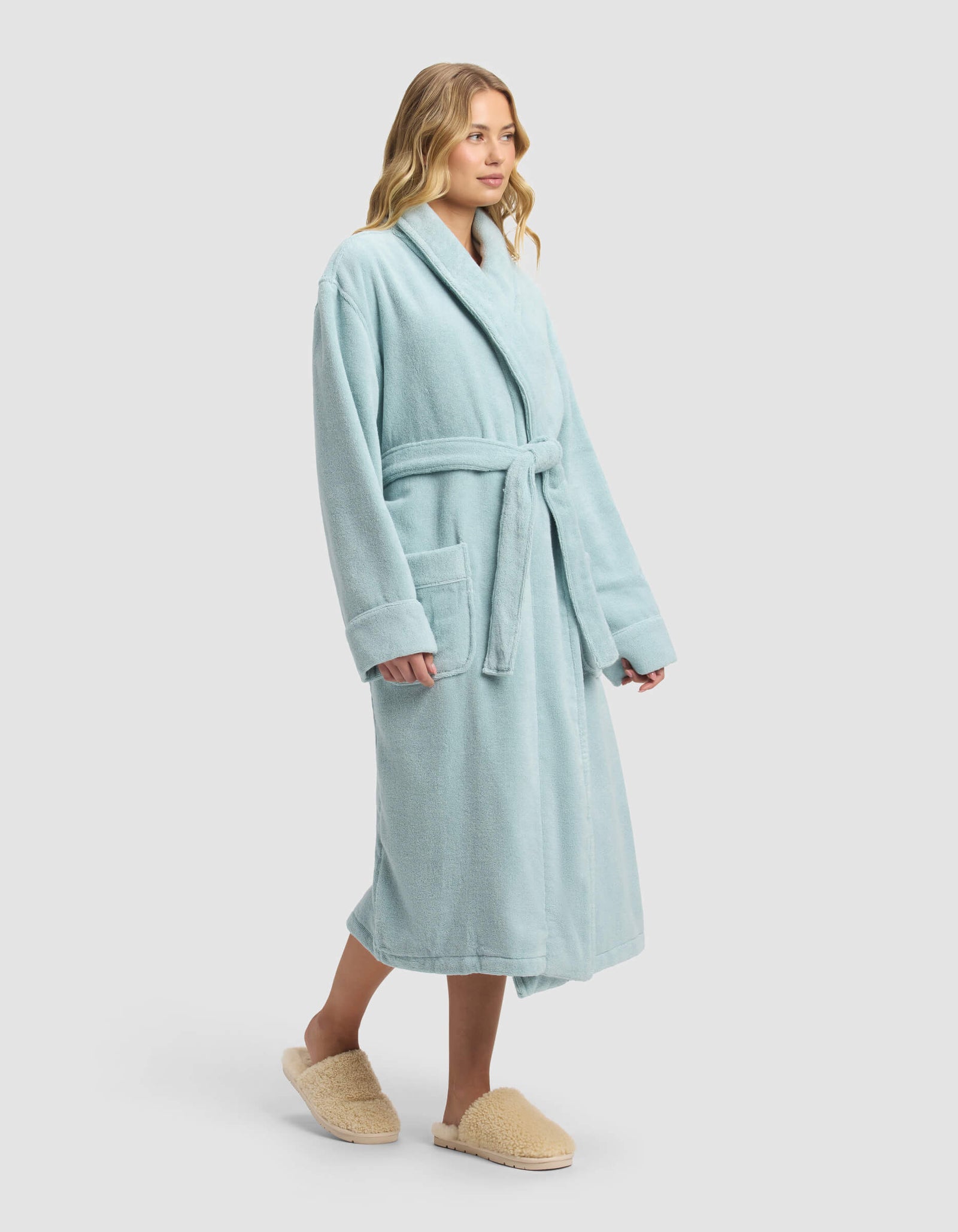 Someone stands against a plain white background wearing the Luxe Bath Robe by Cozy Earth in light blue with a belt and large pockets, paired with beige fuzzy slippers.