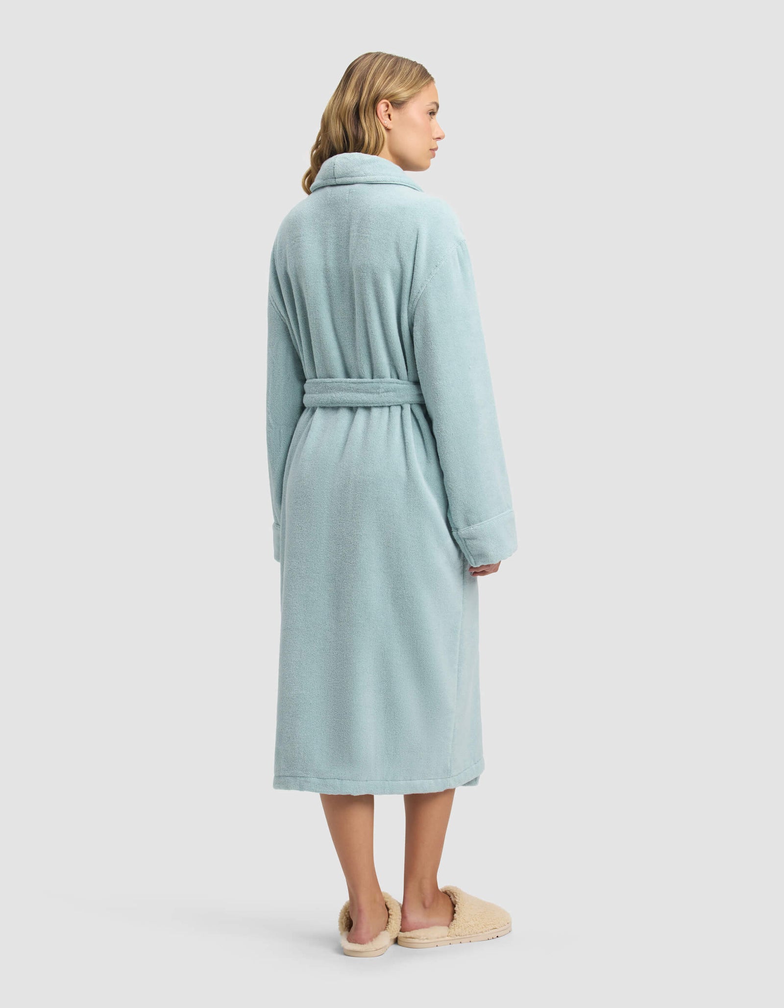 A woman with shoulder-length blonde hair stands facing away against a white background, wearing the Cozy Earth Luxe Bath Robe in light blue with a belt, paired with beige slippers.
