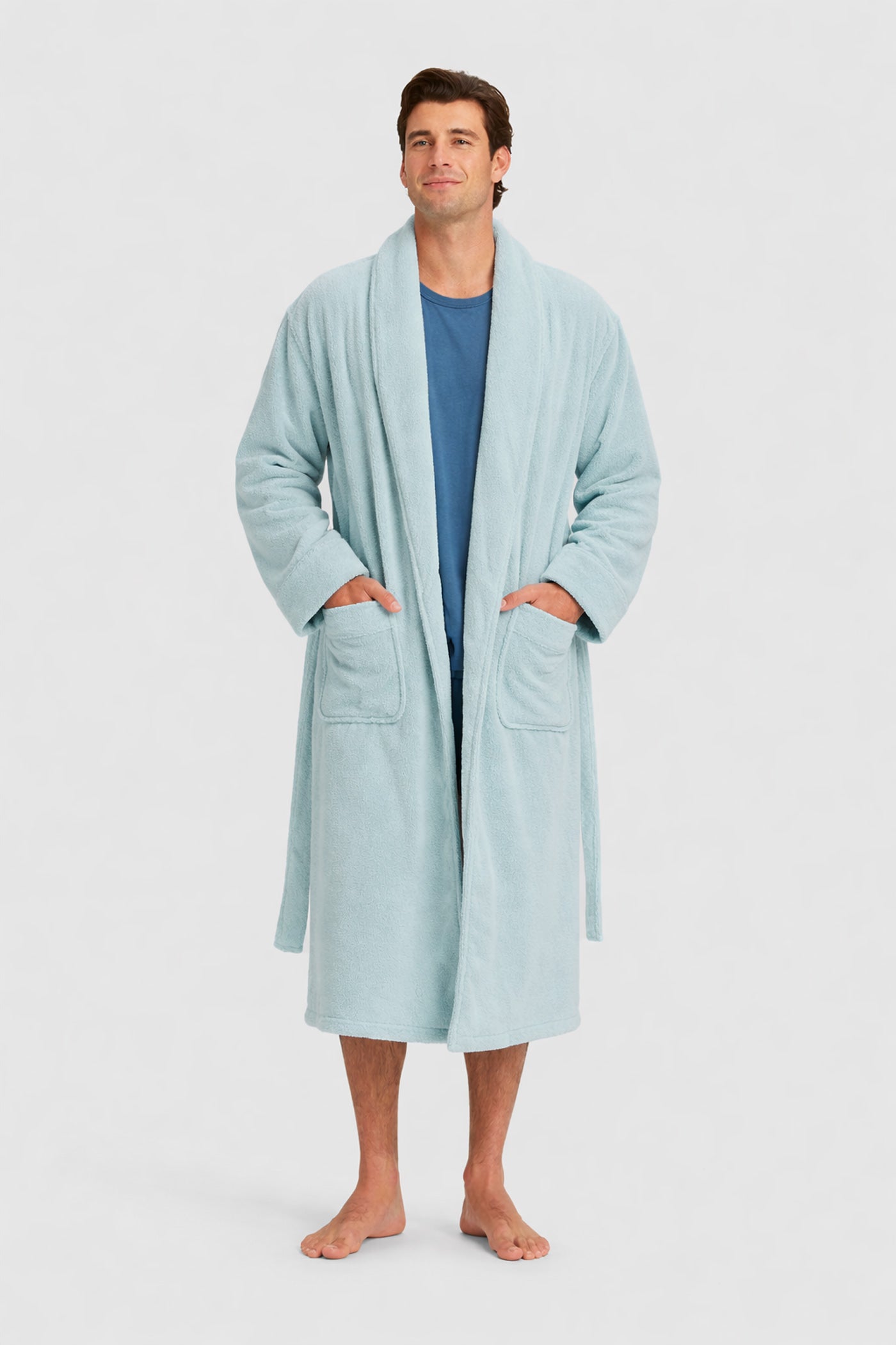 A man stands barefoot in a Luxe Bath Robe by Cozy Earth over a blue shirt, hands in the robe’s pockets, smiling gently against a plain light background.