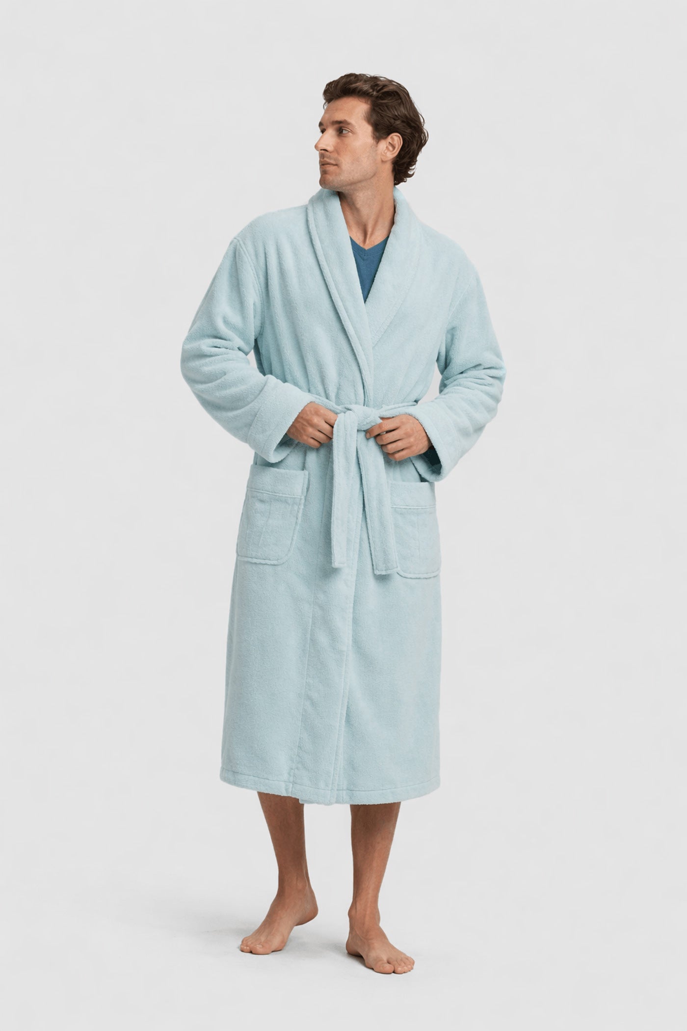 A man stands barefoot in the Luxe Bath Robe by Cozy Earth—a light blue, knee-length robe with a belt and two front pockets—over a blue shirt, looking to his left against a plain light background.